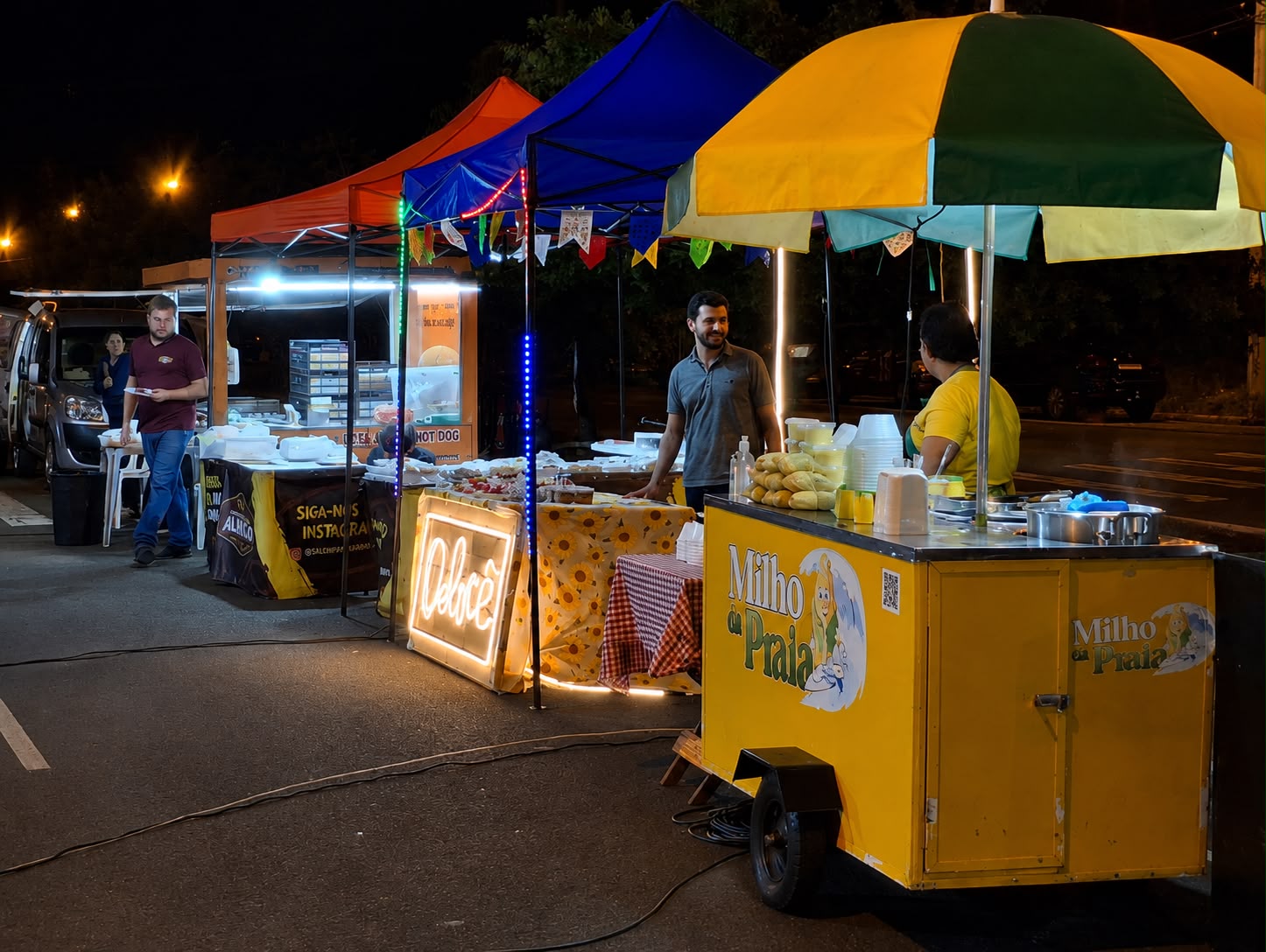 Carrinho do Milho da Praia atendendo em festa junina à noite ao lado de outras barracas